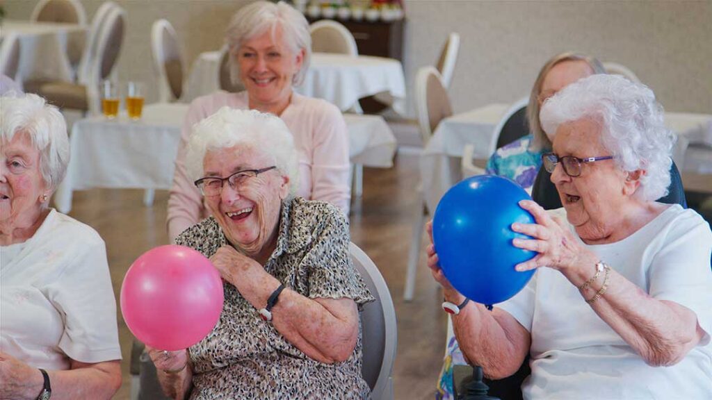 Older adult and family member organize labeled clothing, toiletries, photos, and personal items in a bright assisted living room.